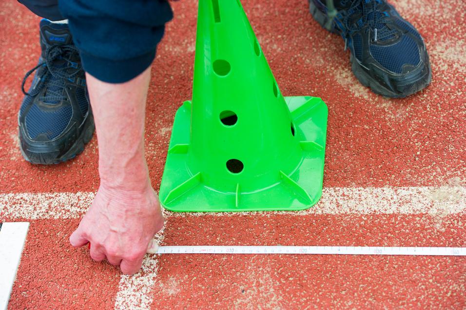 Hand misst mit einem Zollstock die Distanz zu einem grünen Pylonen auf einer roten Sportfläche.